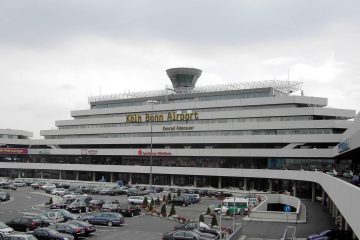 Almanya’da Köln ve Bonn şehirlerine hizmet veren Köln Bonn Havalimanı terminal binası, önünde bekleyen yolcu otobüsleri, taksiler ve arka planda park etmiş uçaklarla birlikte görülüyor.