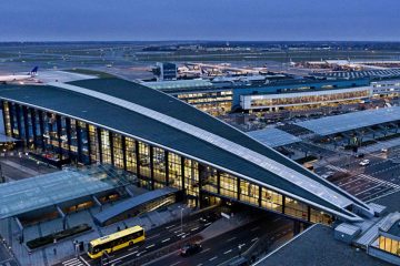 Danimarka’daki Kopenhag Havalimanı terminalinde, geniş cam cepheli modern binanın önünde park etmiş yolcu uçakları, yürüyen yolcular ve apron üzerinde yer hizmetleri araçları görülüyor.