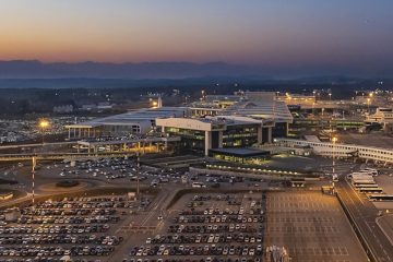 Milano Malpensa Havalimanı pistinde park halinde duran özel jet, arka planda terminal binası ve Milano şehir manzarası ile birlikte görülüyor.